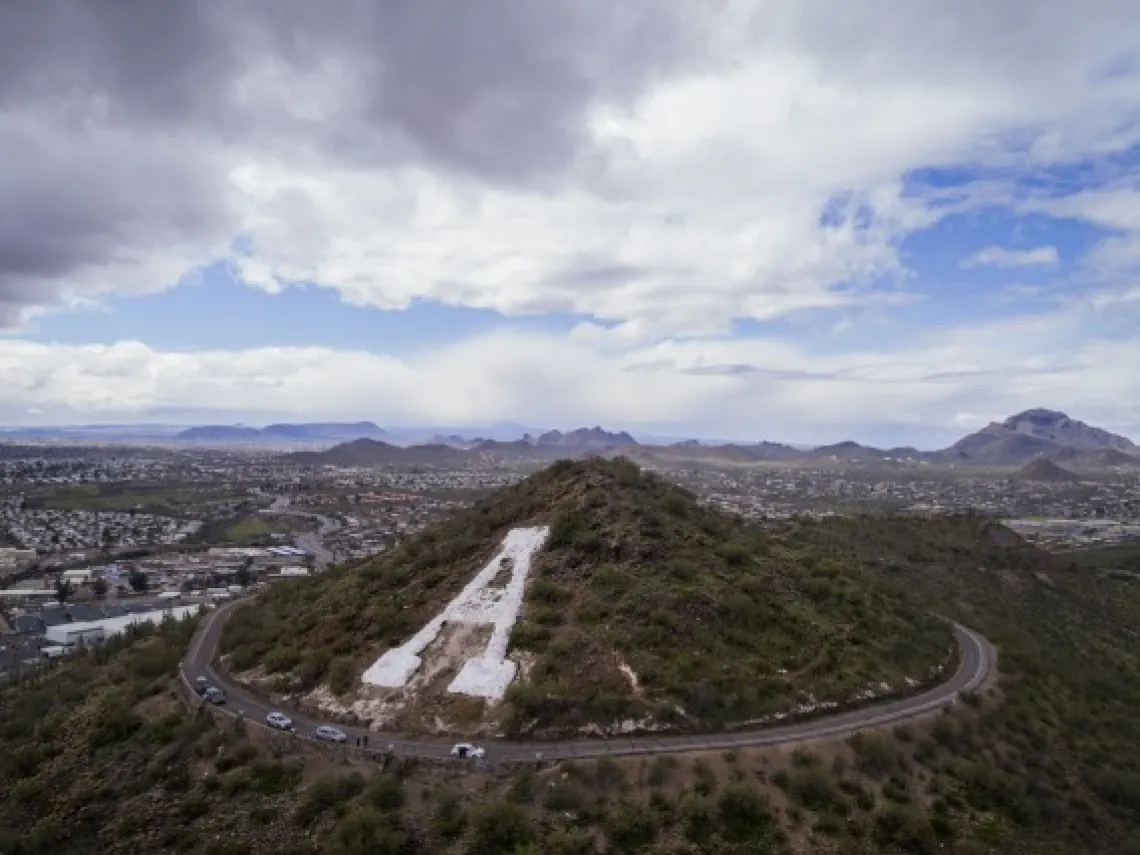 A scenic view of a mountain with a large white-painted 'A' on its slope, commonly known as 'A' Mountain. A winding road circles the base of the hill, with cars visible along the road. The backdrop includes a sprawling cityscape, surrounded by mountain ranges under a cloudy sky, with patches of blue visible.