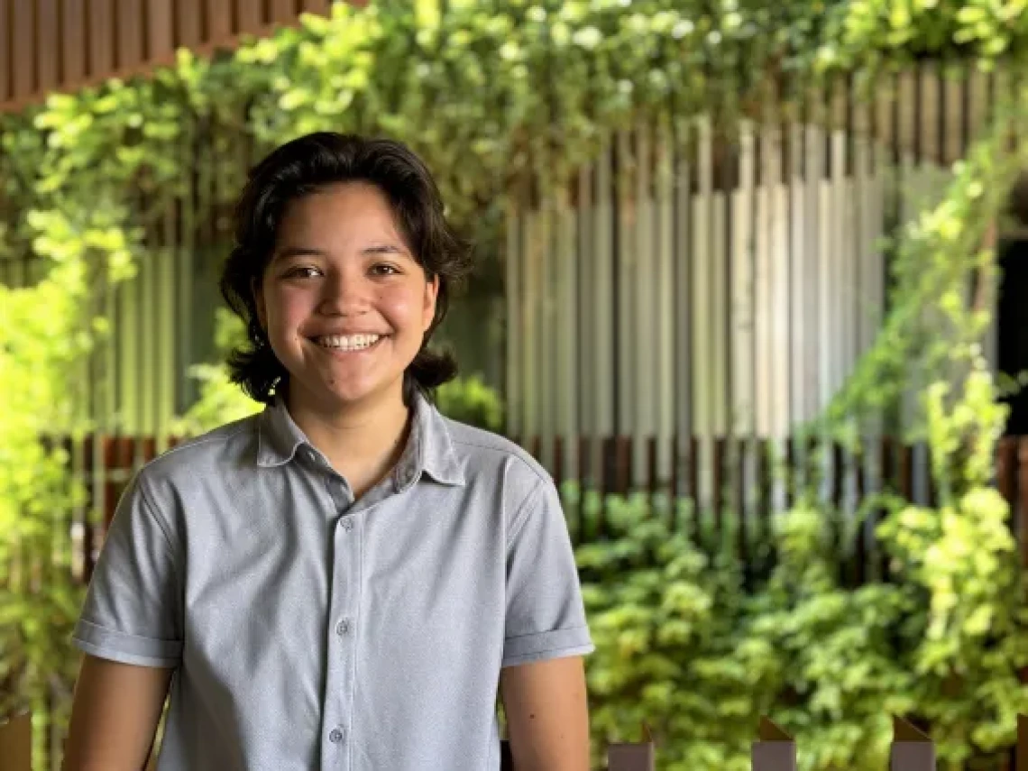 A young hispanic woman with shoulder length hair and is wearing a grey, short sleeve button down shirt and smiling in front of a background of verticle lines mixed with green foliage.