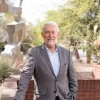 A man with white hair and a beard stands outdoors, leaning against a brick wall with one hand in his pocket. He is wearing a light blue dress shirt and a gray blazer, smiling at the camera.