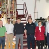 A group of six professionals stands in front of an industrial research setup with metal pipes, wiring, and machinery. The team members wear a mix of business casual and professional attire, with some dressed in polo shirts, blazers, and button-down shirts. A sign in the background reads 'ABSOLUTELY NO STORAGE BELOW,' emphasizing the safety precautions in the high-tech facility.