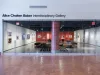 An open space with art on white and red walls and lounge seating sits behind a wall of glass doors set into a white wall. Above the doors reads "Alice Chainten Bker Interdisciplinary Gallery"